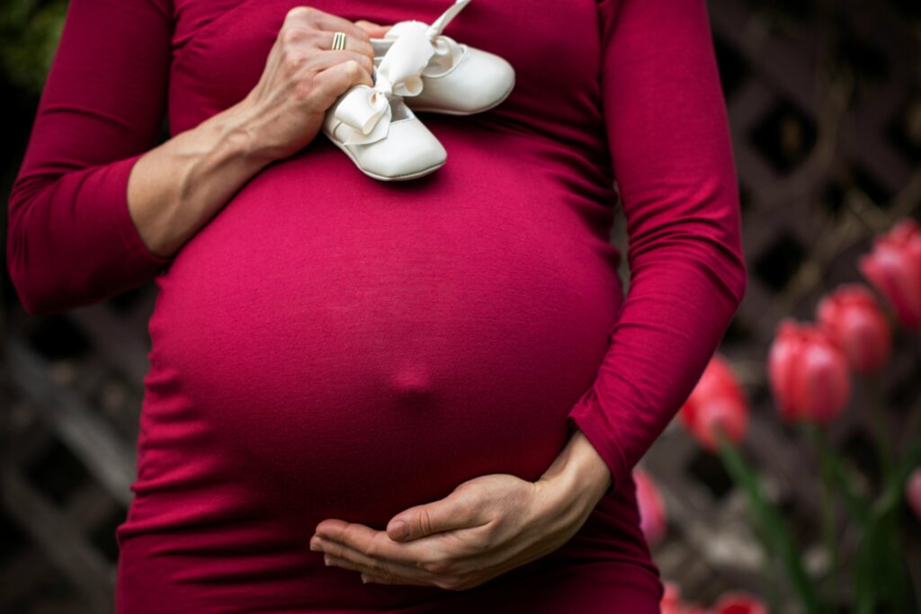 Pregnant woman holding baby shoes on belly in garden setting.